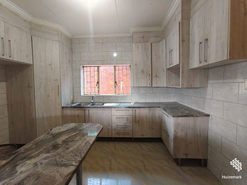 A clean and modern kitchen featuring light wood-grain cabinetry, stone-look countertops, a double sink, and neutral wall tiling.