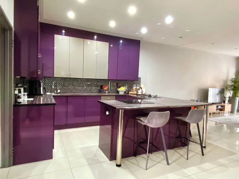 Modern open-plan kitchen featuring high-gloss purple cabinetry, a central island with integrated hob, and a mosaic tile backsplash, seamlessly flowing into the living area.