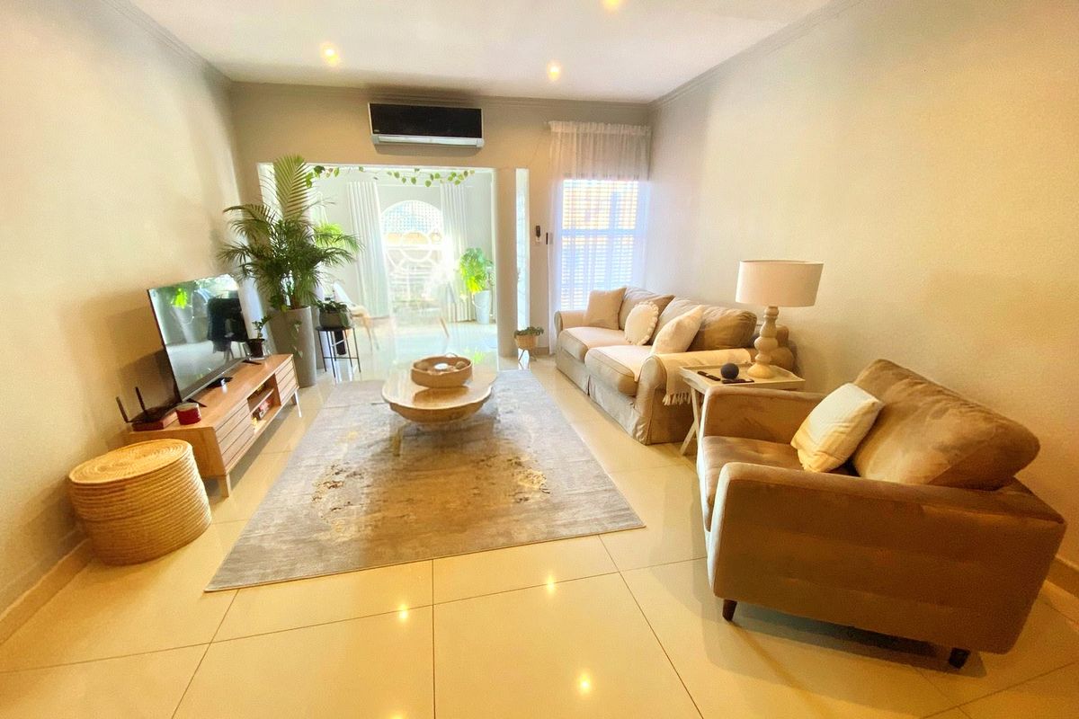 A well-lit living room featuring tiled flooring, recessed downlighting, and a wall-mounted split-system air conditioner, leading to an adjacent sunlit area.