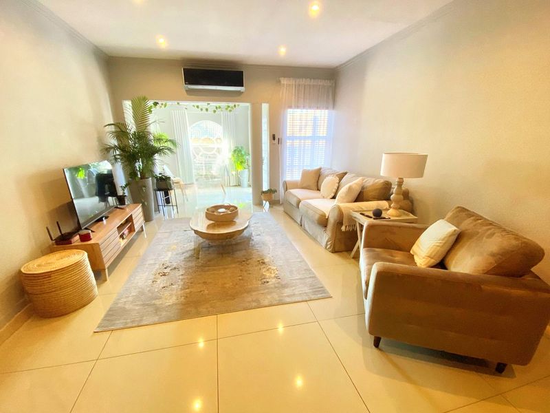 A well-lit living room featuring tiled flooring, recessed downlighting, and a wall-mounted split-system air conditioner, leading to an adjacent sunlit area.