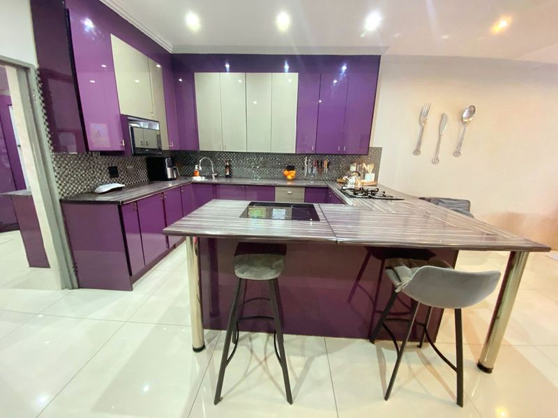 Modern kitchen featuring high-gloss purple and cream cabinetry, a central island with integrated hob, mosaic tile backsplash, and polished tiled flooring.