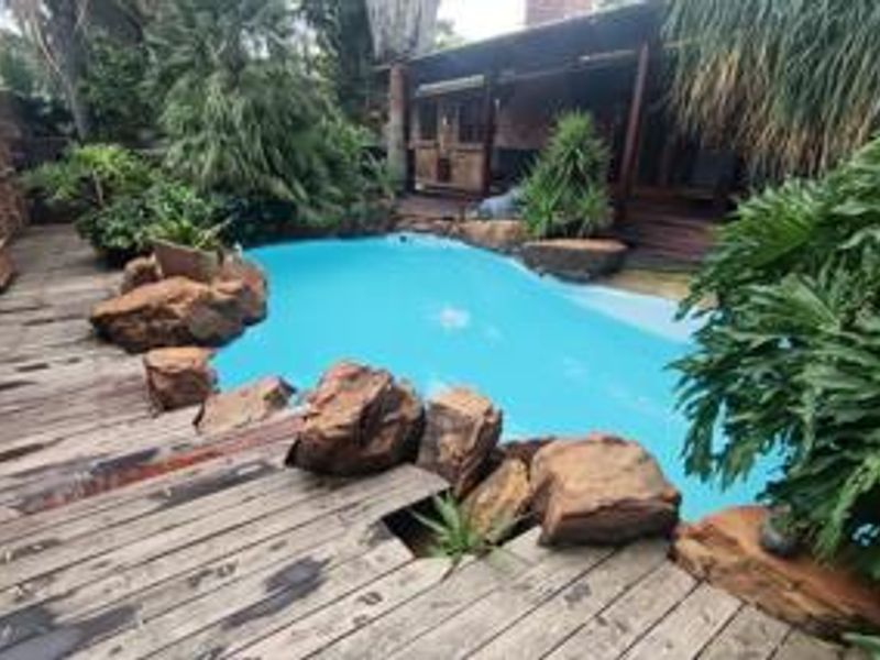 Outdoor pool area featuring a swimming pool surrounded by natural rock landscaping and timber decking, adjacent to a covered patio space.