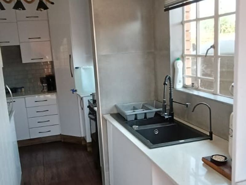 A functional scullery area featuring modern white cabinetry, stone countertops, a dark undermount sink, and wood-look tiled flooring, leading into the main kitchen.