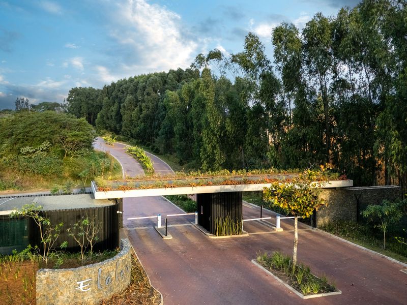 Aerial view of a modern residential estate entrance featuring a landscaped security gatehouse, paved driveway, and established perimeter trees.