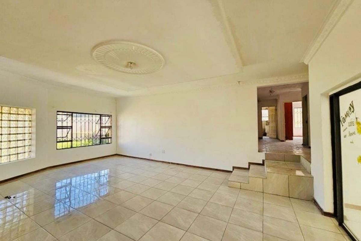 Spacious, empty living area featuring tiled flooring, decorative ceiling rose, and a raised platform section with access to an adjoining hallway.