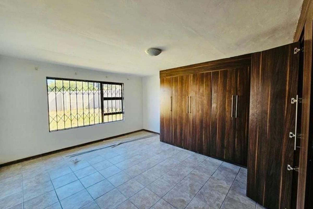 A spacious, empty bedroom featuring tiled flooring, built-in wooden cupboards, and a window fitted with security bars.