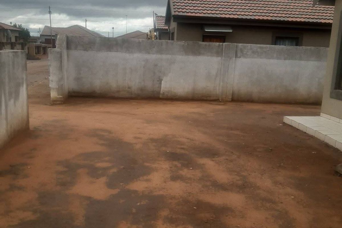 A view of the rear yard featuring a concrete boundary wall and tiled roof structure, with an unpaved ground surface.
