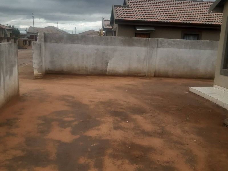 A view of the rear yard featuring a concrete boundary wall and tiled roof structure, with an unpaved ground surface.