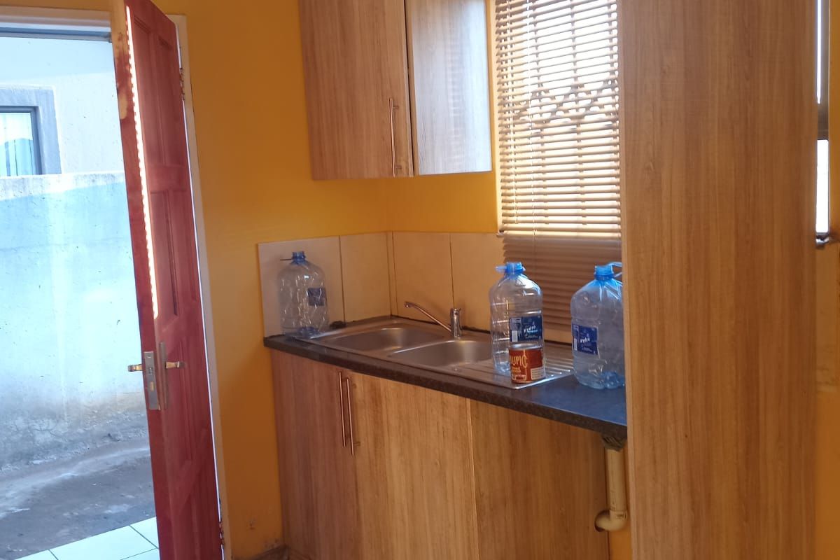 A compact kitchen area featuring light wood-look cabinetry, a stainless steel sink, tiled flooring, and a tiled splashback.