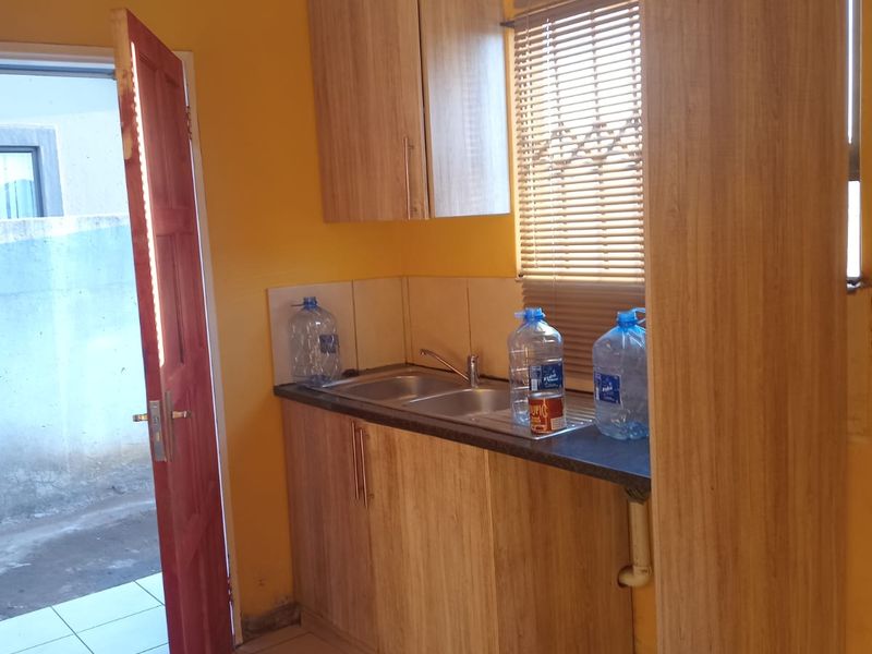A compact kitchen area featuring light wood-look cabinetry, a stainless steel sink, tiled flooring, and a tiled splashback.