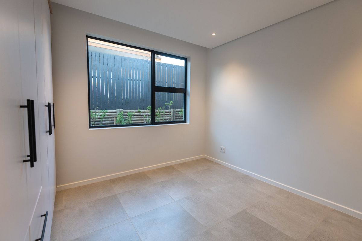 A bright, modern, and empty bedroom featuring neutral tiled flooring, built-in cupboards, and a large window with a view of the rear garden fence.