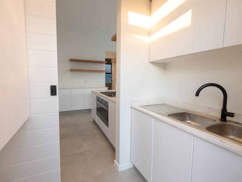 Modern kitchen featuring clean white cabinetry, stone countertops, integrated appliances, and floating timber shelving, finished with contemporary black fixtures and neutral floor tiles.
