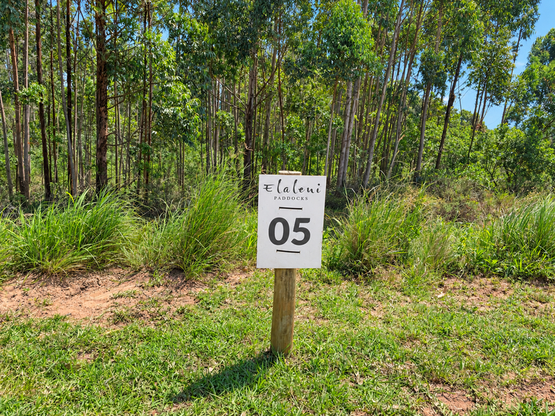 A vacant residential plot marked with a property identification sign, featuring natural vegetation and mature trees in a peaceful setting.