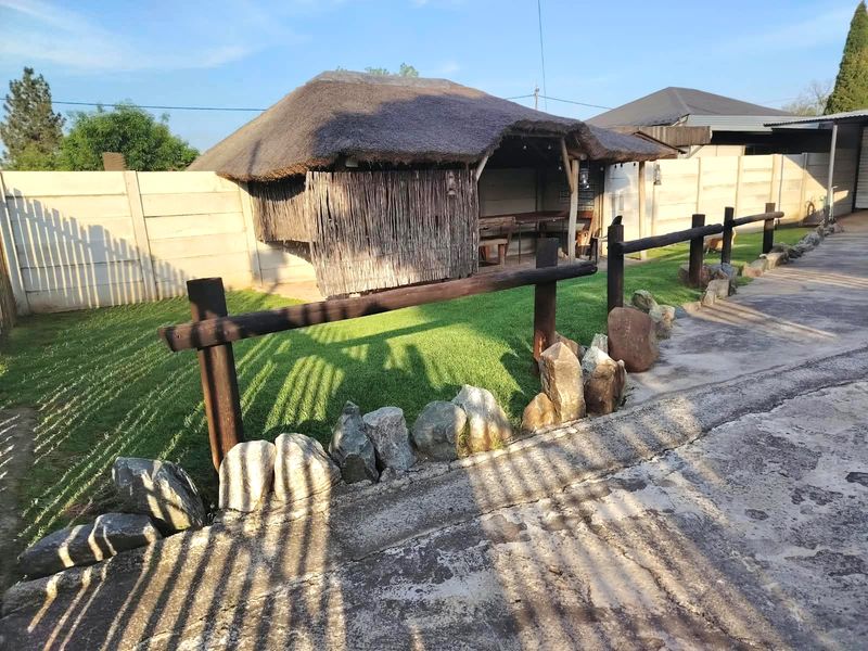 Outdoor entertainment area featuring a thatch-roofed lapa, enclosed by precast concrete walls and timber fencing, set within a landscaped garden with stone borders.