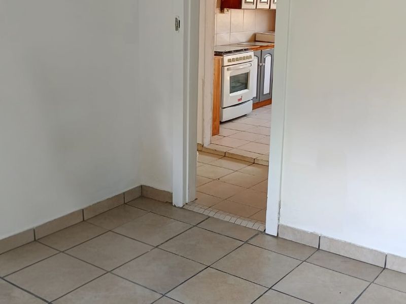 An empty living area with ceramic floor tiles, featuring a doorway leading into a kitchen with built-in cabinetry and a stove.