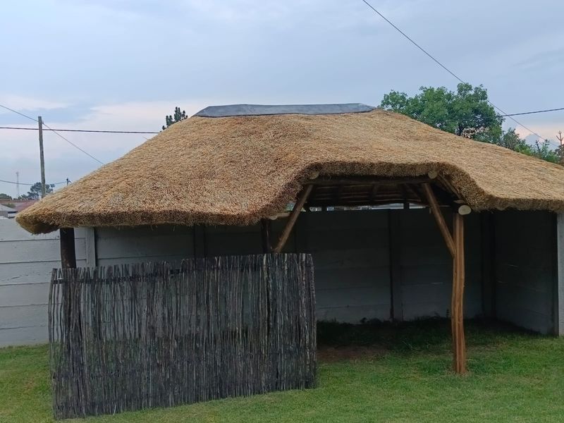 An outdoor thatched lapa structure featuring timber support posts and a precast concrete boundary wall, set within a garden area.