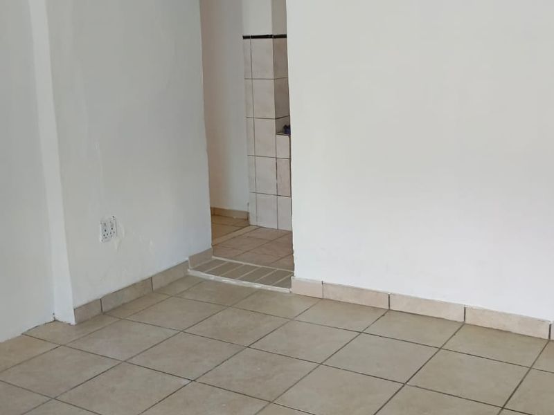 An empty living area featuring neutral ceramic floor tiles and plain white walls, leading into an adjacent tiled utility or kitchen space.