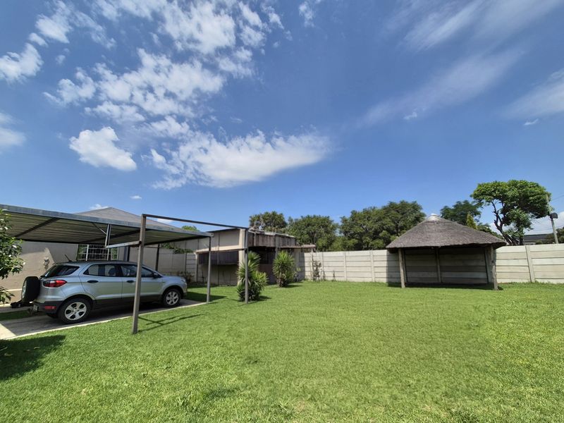 Spacious rear garden featuring a paved driveway, metal carport structure, and a traditional thatched lapa, enclosed by a precast concrete boundary wall.