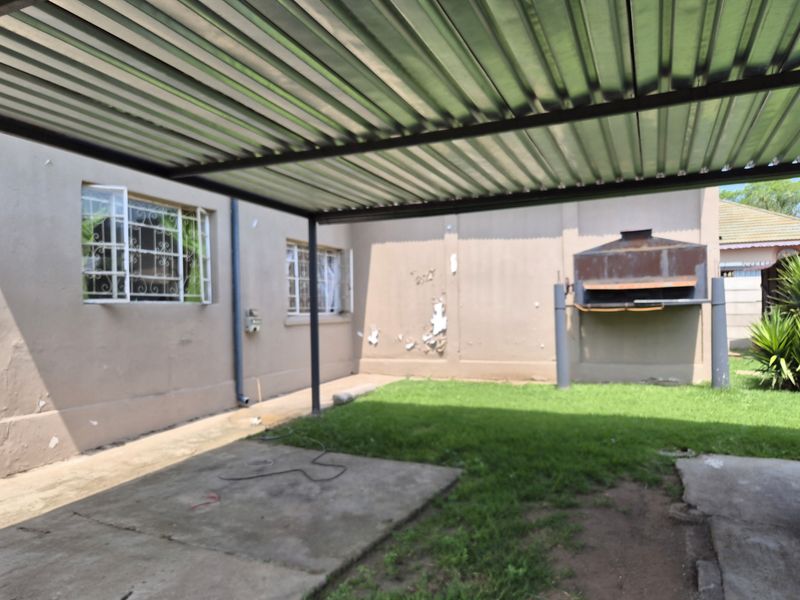Covered outdoor patio area featuring a built-in braai, corrugated metal roofing, and security-barred windows, requiring some exterior maintenance and paint touch-ups.