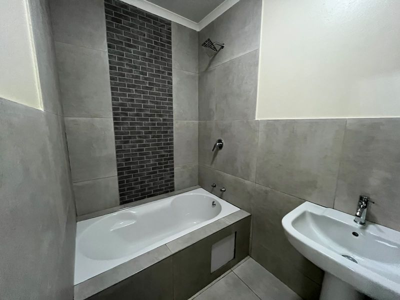 A modern bathroom featuring a built-in bathtub with a shower head, pedestal basin, and contemporary grey wall tiles with a decorative brick-style feature panel.