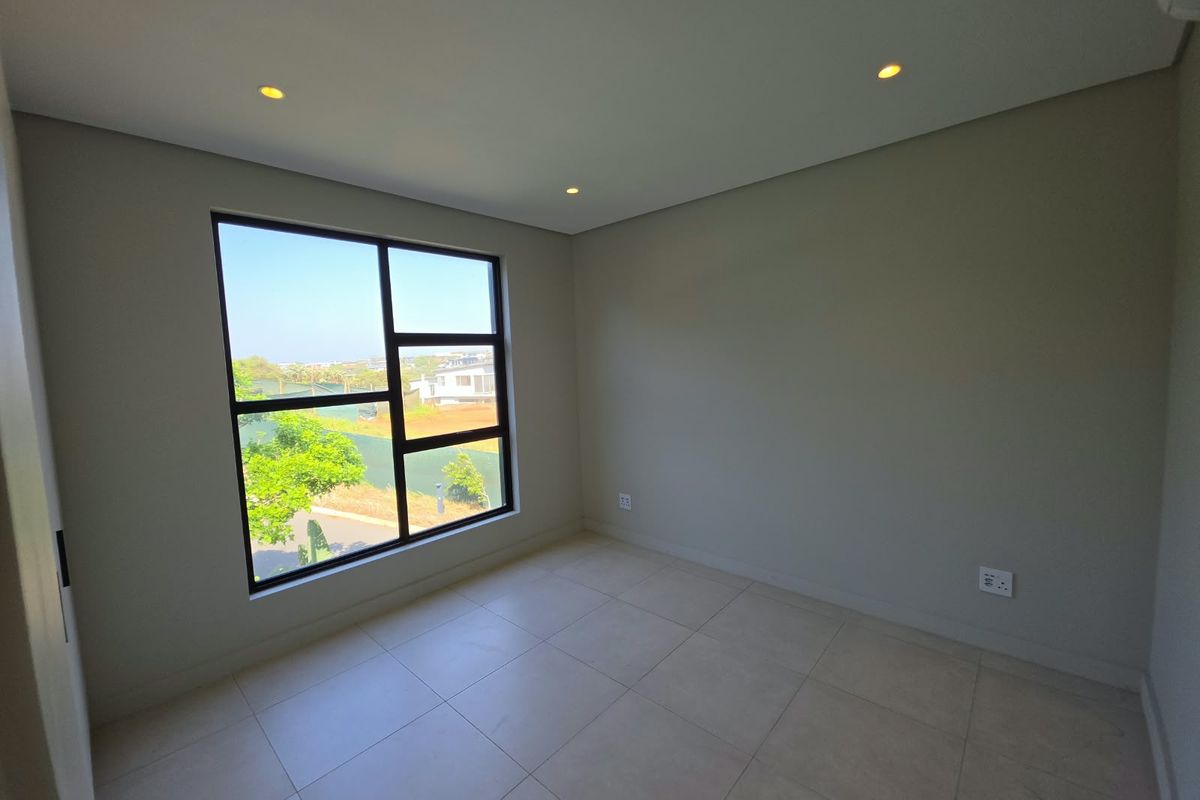 A bright, empty bedroom featuring modern tiled flooring, recessed downlighting, and a large window with views of the surrounding area.