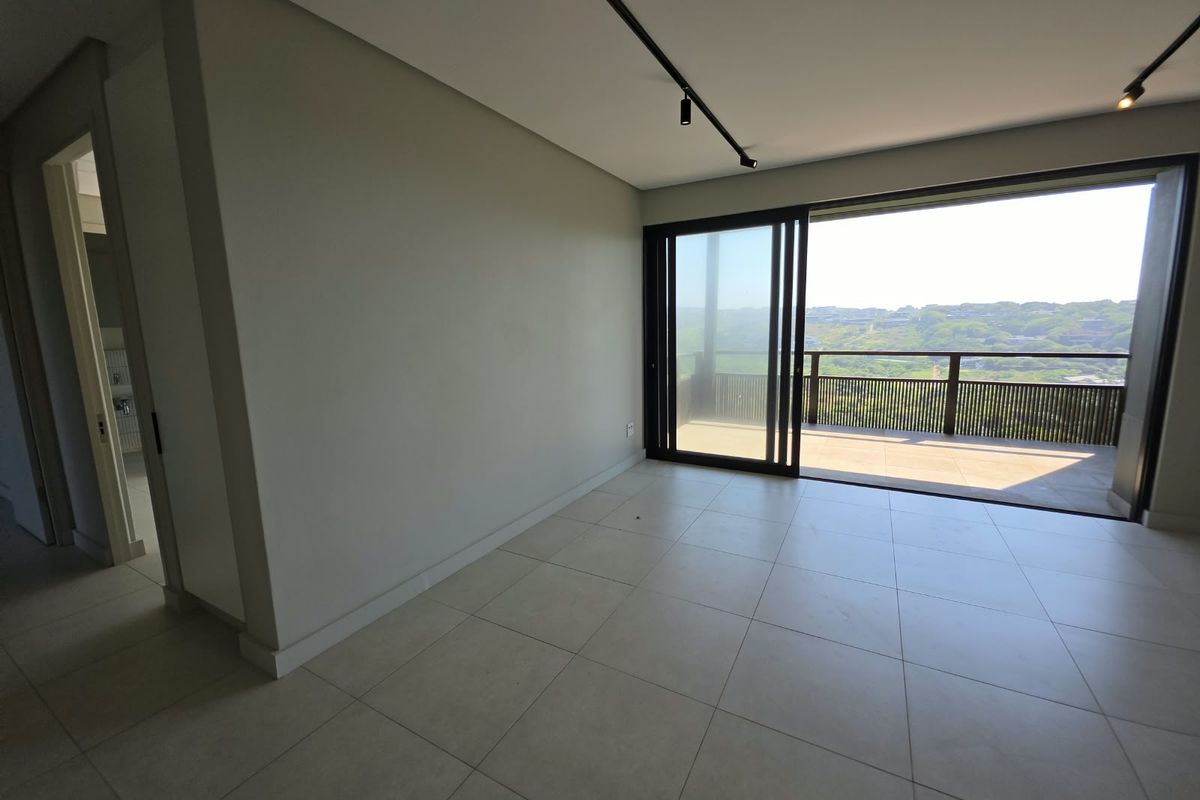 A bright, modern, and empty living area featuring large format floor tiles, track lighting, and expansive sliding glass doors leading to a balcony with scenic views.