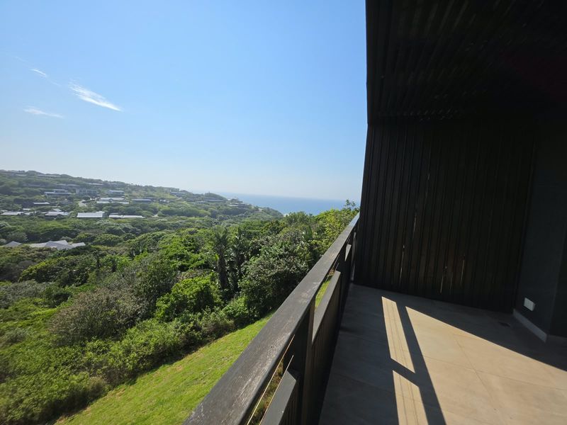A spacious covered balcony featuring timber cladding and a balustrade, offering expansive views of the surrounding coastal landscape and greenery.