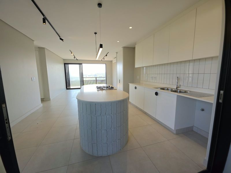 Modern open-plan kitchen featuring a central island with a built-in hob, sleek cabinetry, and tiled backsplash, flowing into a bright living area with balcony access.