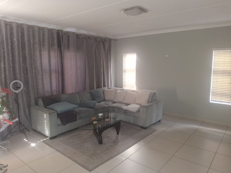 A spacious living room featuring neutral tiled flooring, large windows with venetian blinds, and a simple ceiling light fixture.