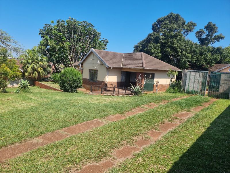 A detached single-story house featuring a tiled roof and brick facade, set on a spacious plot with a grass driveway and established garden.