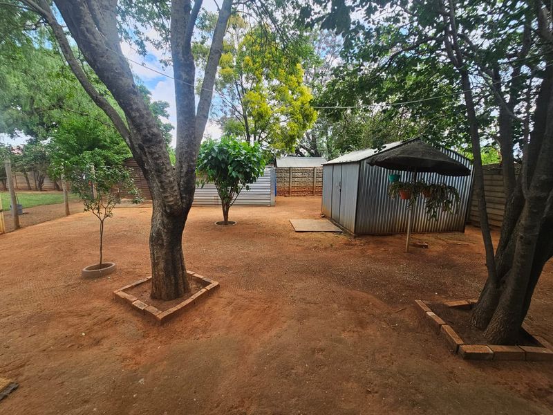 Spacious backyard featuring mature trees, brick-edged garden beds, a metal storage shed, and a precast concrete boundary wall.