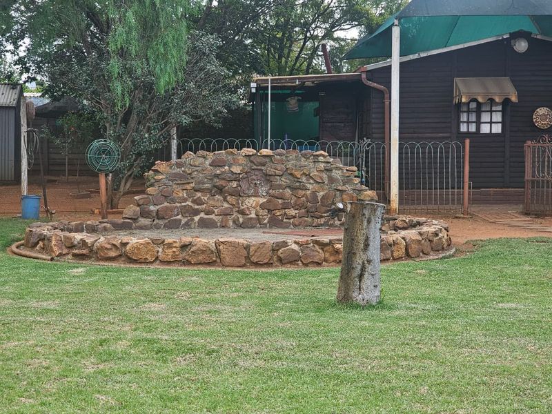 A garden area featuring a stone water feature, lawn, and metal fencing, adjacent to a timber-clad structure with a shade sail.