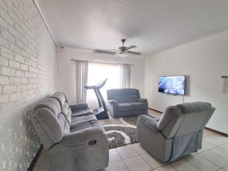 A living room featuring tiled flooring, an exposed white brick feature wall, and a ceiling fan, providing a functional and comfortable living space.