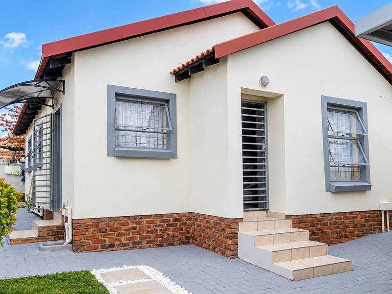 A well-maintained front elevation featuring a tiled roof, brick plinth detailing, secure window and door grilles, and a neat paved driveway with carport.