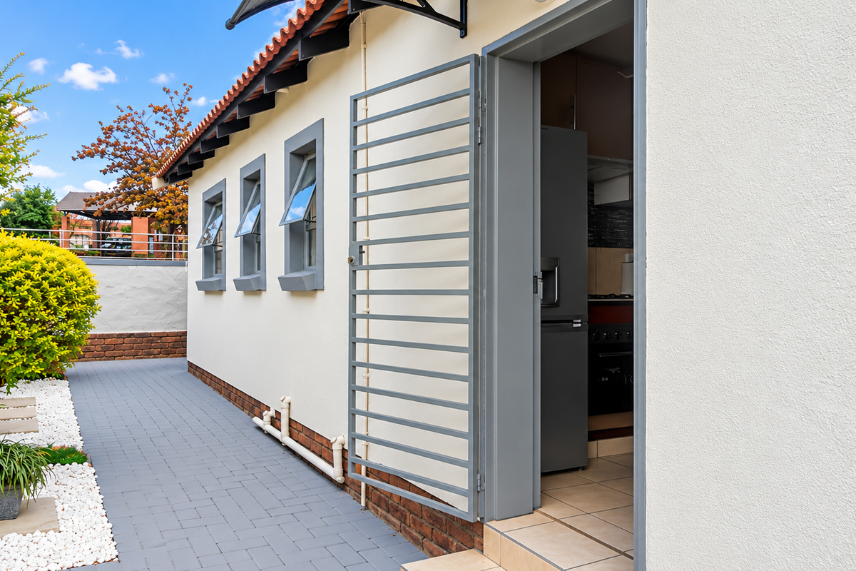 A well-maintained rear exterior view featuring a paved walkway, security gate, and access to the interior kitchen area.