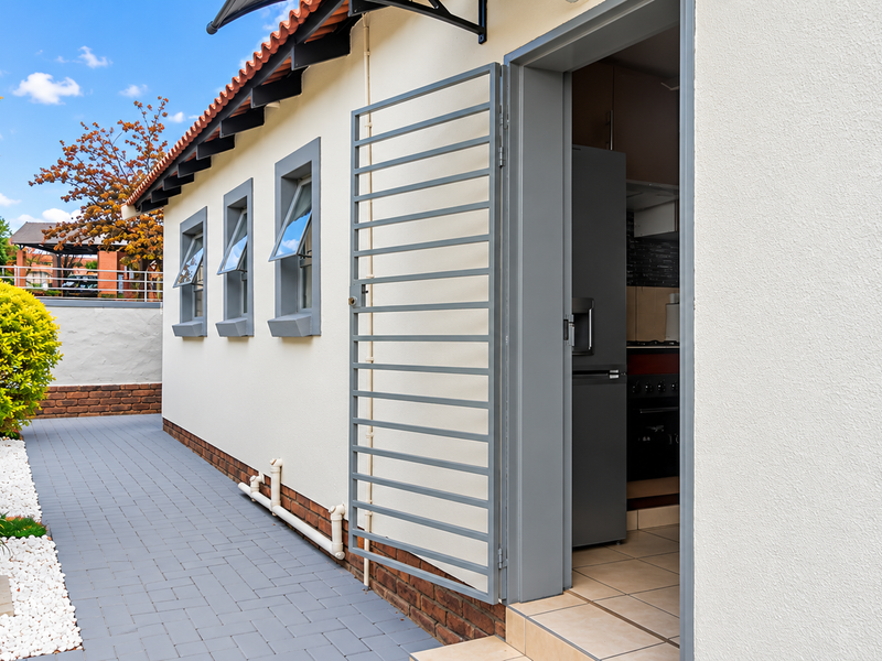 A well-maintained rear exterior view featuring a paved walkway, security gate, and access to the interior kitchen area.