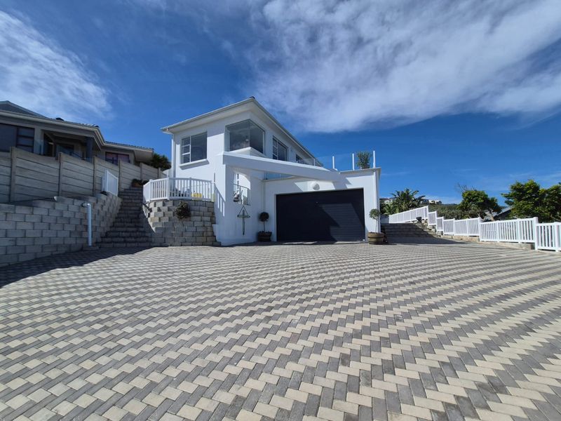 Modern front elevation featuring a paved driveway, double garage, and elevated terrace with glass balustrades, set against a clear blue sky.
