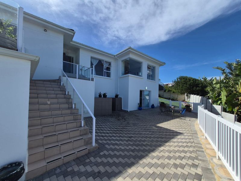 A well-maintained rear exterior featuring a paved patio area, external staircase with glass balustrades, and a white picket fence bordering the garden.