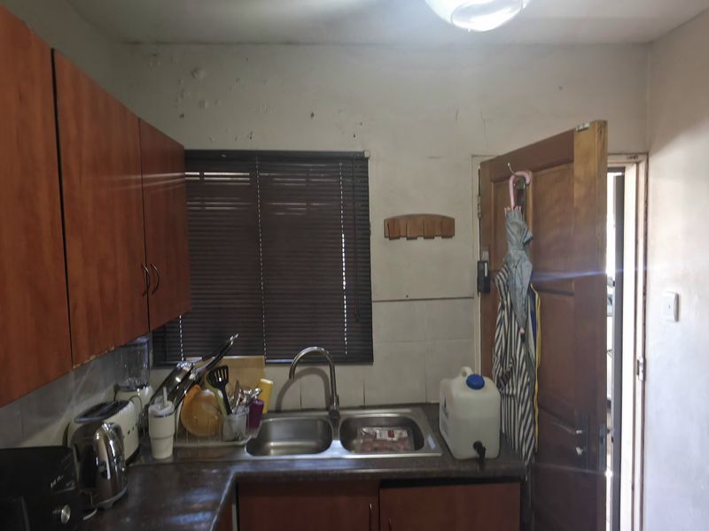 A compact kitchen featuring laminate cabinetry, a double bowl stainless steel sink, and a window with venetian blinds, requiring cosmetic updates and maintenance.