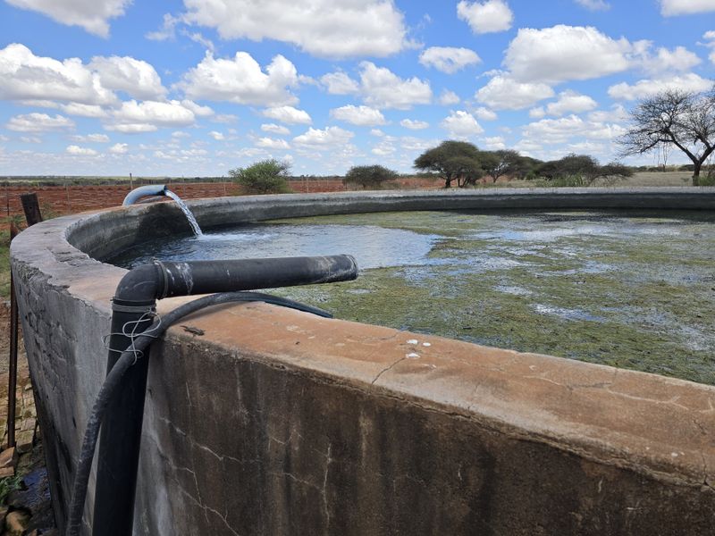 A large circular concrete water reservoir on the farm, featuring an active water inlet pipe and visible surface algae.