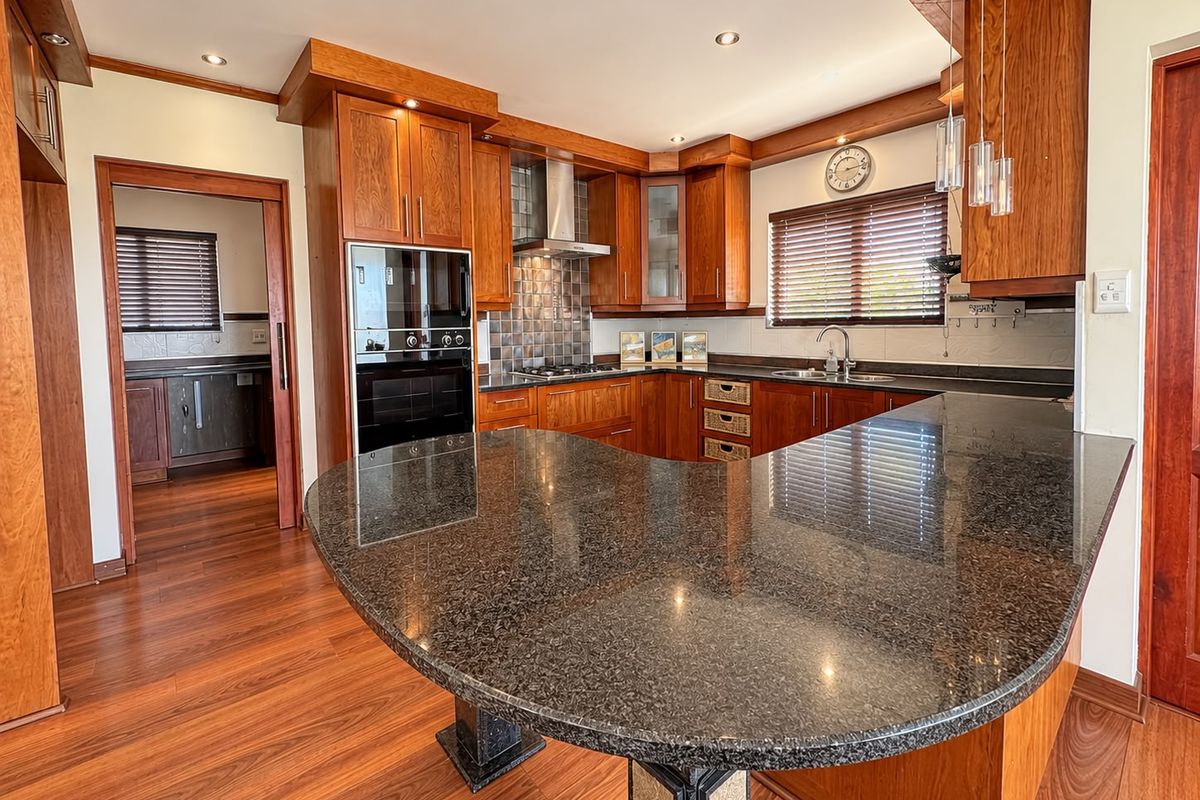 A well-maintained kitchen featuring rich wooden cabinetry, granite countertops, a breakfast bar, and integrated appliances, with a seamless flow into the adjacent scullery area.