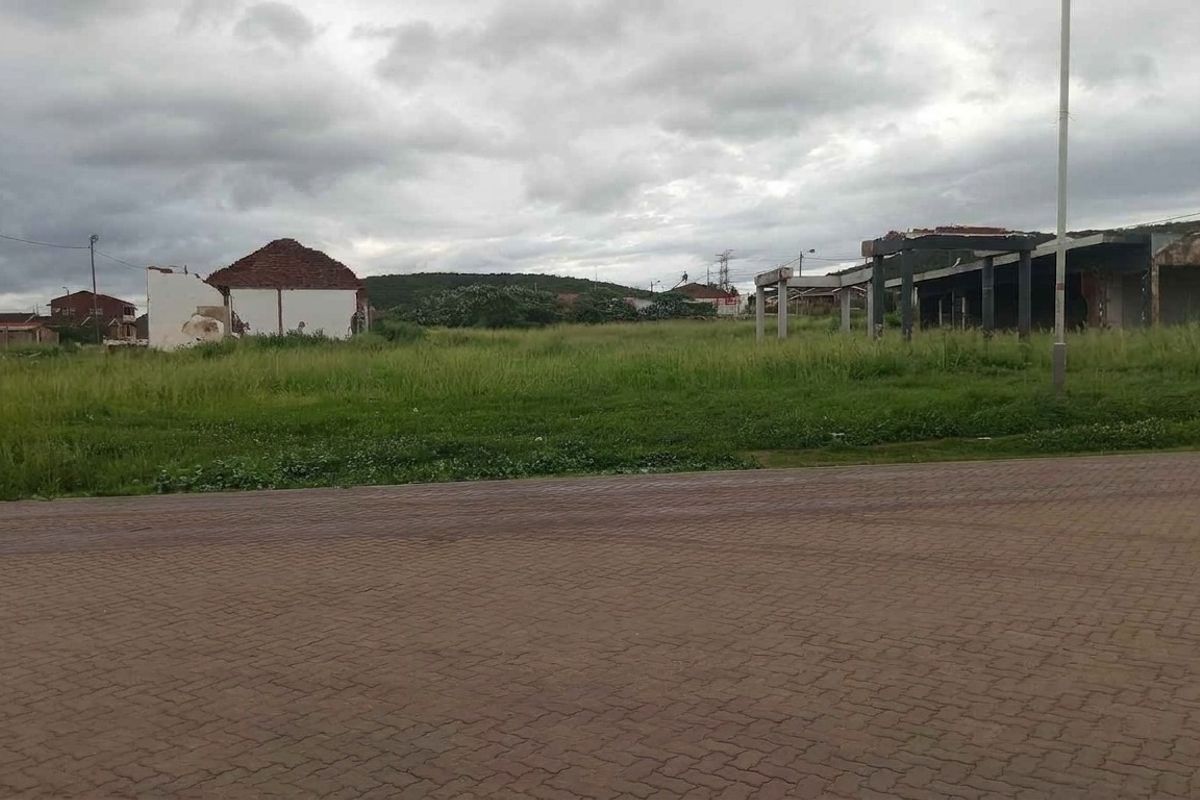 A vacant residential plot featuring overgrown grass and the remains of dilapidated structures, situated along a paved road.