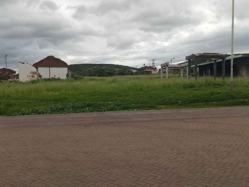 A vacant residential plot featuring overgrown grass and the remains of dilapidated structures, situated along a paved road.