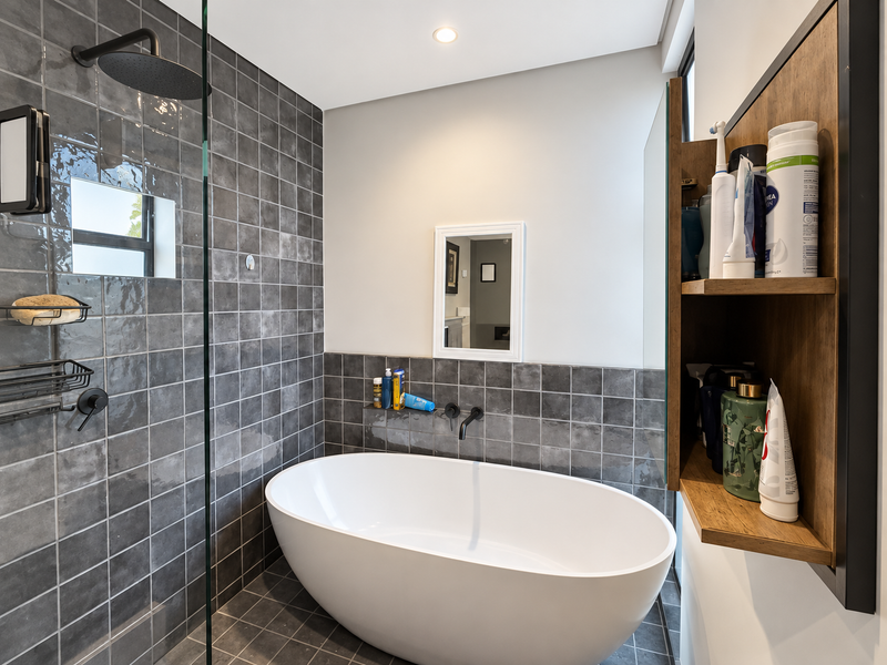 Modern bathroom featuring a sleek freestanding bathtub, wall-mounted fixtures, a glass-enclosed rain shower, and stylish grey tiled finishes with integrated shelving.