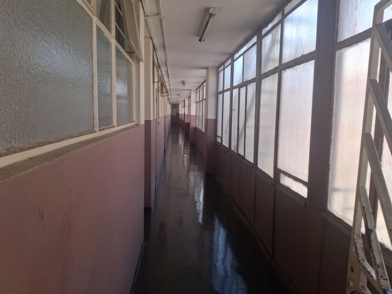 A long, enclosed apartment hallway featuring metal-framed windows with wired glass and fluorescent lighting, showing signs of wear and dated finishes.