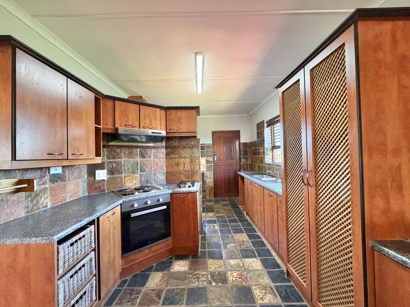 A functional kitchen featuring warm-toned wooden cabinetry, stone tile flooring, a built-in oven with hob, and ample storage space with louvered pantry doors.