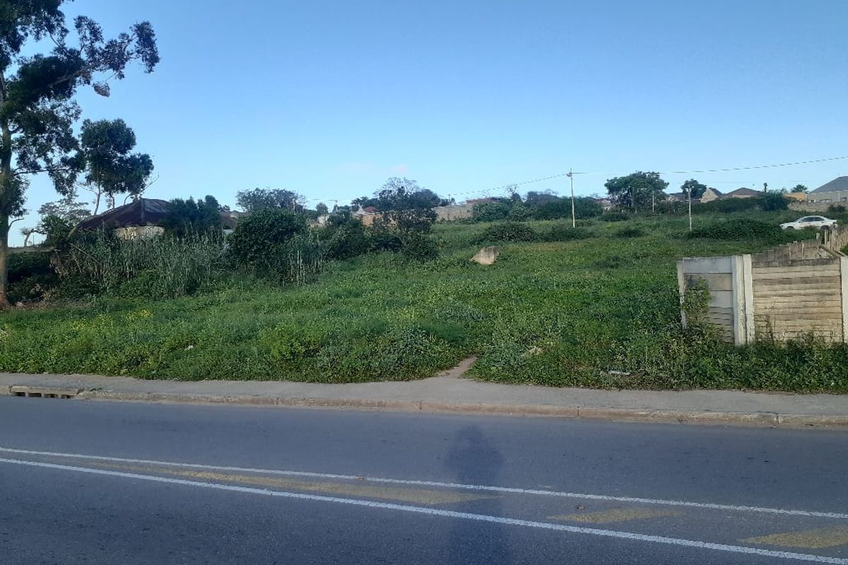 A vacant residential plot featuring a concrete boundary wall, accessible from an asphalt road with an adjacent paved sidewalk.