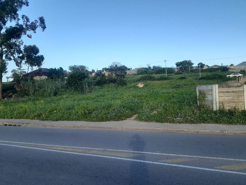 A vacant residential plot featuring a concrete boundary wall, accessible from an asphalt road with an adjacent paved sidewalk.