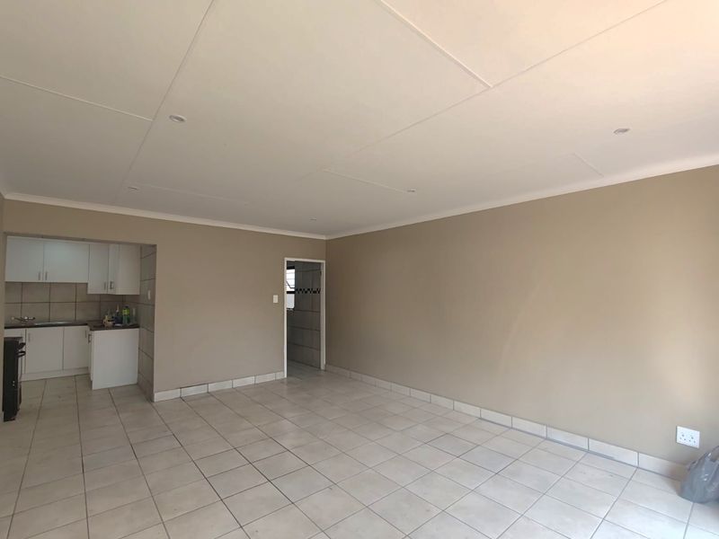 An open-plan living area featuring neutral wall tones, tiled flooring, and a compact kitchen with built-in cabinetry and sink.