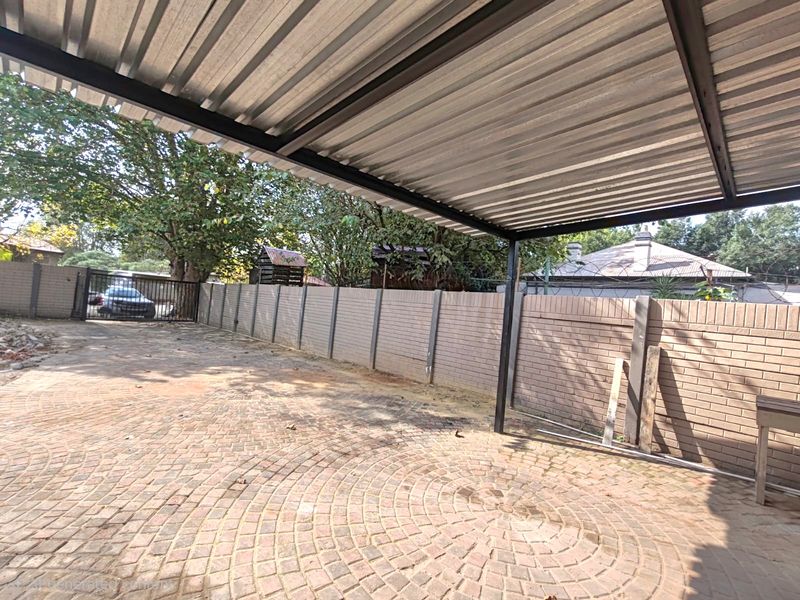 An open-sided carport structure featuring a corrugated metal roof, paved driveway, and secure brick boundary walling.